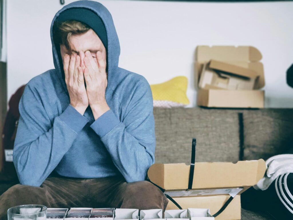 Man in a blue hoodie covers his face with his hands, overwhelmed after a relapse, sitting among cardboard boxes at home.